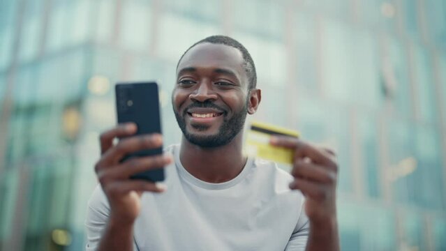 African american man paying online on smartphone adding bank card number standing on street. Smiling guy buying online in phone. Shopping, urban lifestyle, buy in internet, mobile payment concept.