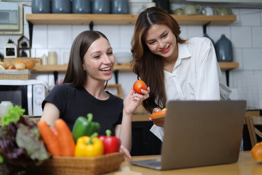 Two young women in the kitchen practicing cooking are looking at recipes on their laptops.