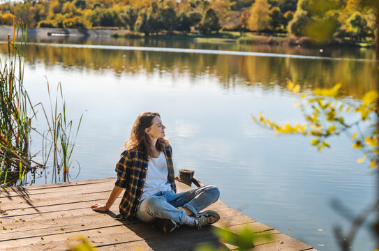 Young woman relaxing sitting on a wooden pier on a pond enjoying autumn nature