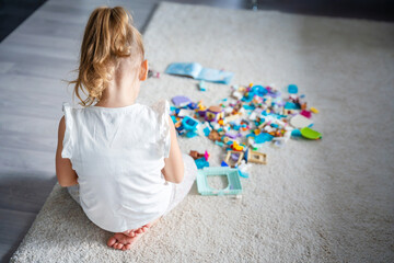View from the back of little girl playing with small constructor toy on floor in home, educational...