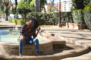 Young woman, beautiful and black with afro hair, with flower shirt, jeans and boots, sitting in a fountain playing with her hair. Concept beauty, relaxation, hairstyles.