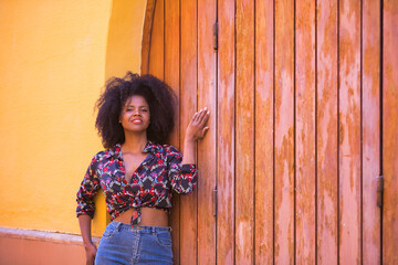 Fototapeta premium Young woman, beautiful and black with afro hair, with a floral shirt leaning on a wooden board door, quiet, happy and smiling. Concept beauty, tranquility, happiness.