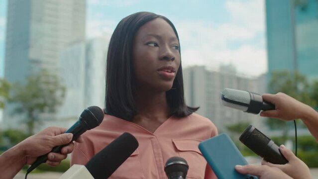 Medium close up of young female black journalist speaking into microphones while giving interview to journalists at street