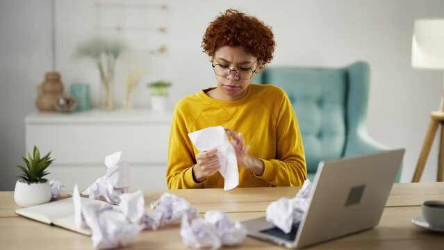 African american girl student writing educational project on paper sheets crumpling bad ones sitting at table on workplace at home. Curly creative woman working on documents. Education, study, learn.
