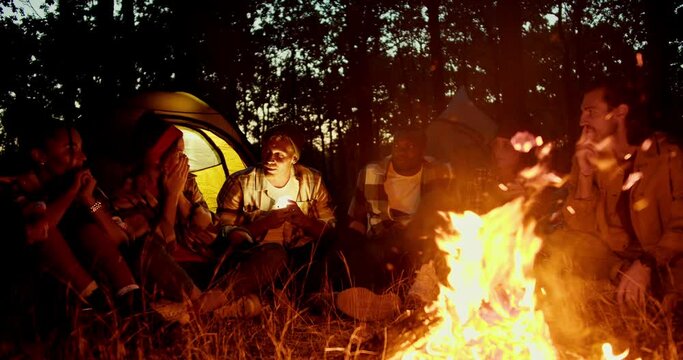 Evening cozy communication near the fire, a small group sits near the fire and tells scary stories against the backdrop of the floor and the evening autumn forest. A man in a hat shines a flashlight