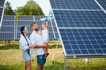 A wide shot of a happy family standing together and smiling at camera with a large solar panel in background