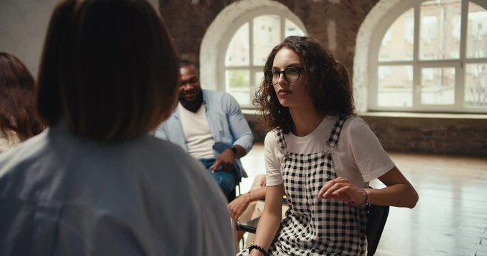 A brunette girl with curly hair in glasses and a checkered jumpsuit communicates in pairs during group therapy in a brown hall