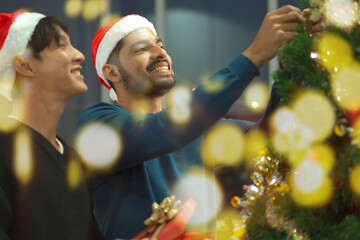Lovely gay couple enjoy decorating a Christmas tree together in a living room with beautiful festive light bulbs and presents. LGBTQ gay couple celebrating a Christmas Eve and New Year festival. - obrazy, fototapety, plakaty