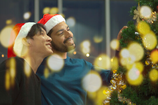 Lovely gay couple enjoy decorating a Christmas tree together in a living room with beautiful festive light bulbs and presents. LGBTQ gay couple celebrating a Christmas Eve and New Year festival. - Powered by Adobe