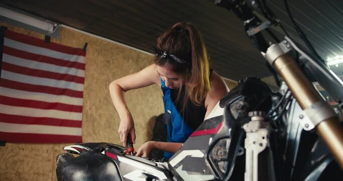 close-up shot: a girl auto mechanic repairs a motorcycle in her workshop with a screwdriver against the background of the US flag and tools in the garage