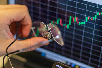 Close up shot of the hand with glasses next to the computer screen with trading bar charts. Finance