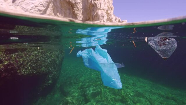 Split level, Plastic bag and other debris floats on surface of water near roks in suny day, Mediterranean Sea, Slow motion. Global plastic pollution of seas and oceans