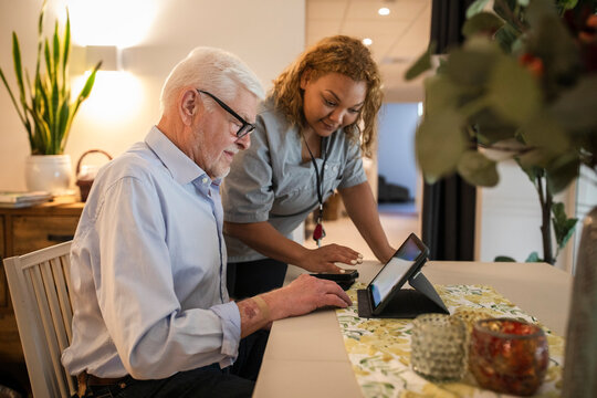 Young female caregiver assisting senior man in using digital tablet at retirement home
