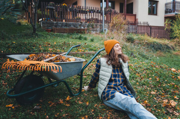 Young happy caucasian woman collecting autumn leaves in the backyard of a country house using a rake and cart