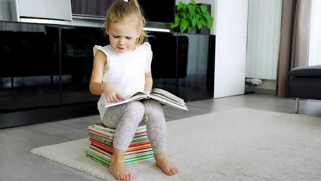 Cute Little girl is sitting on stack of children's books and leafing through a book with fairy tales