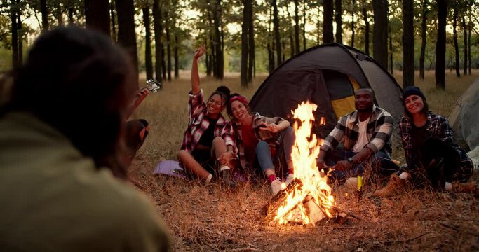 A brunette man in a green jacket takes photographs of group hikers in checkered shirts near a fire. A man plays a guitar and people pose in the frame near a tent in a green and yellow autumn forest