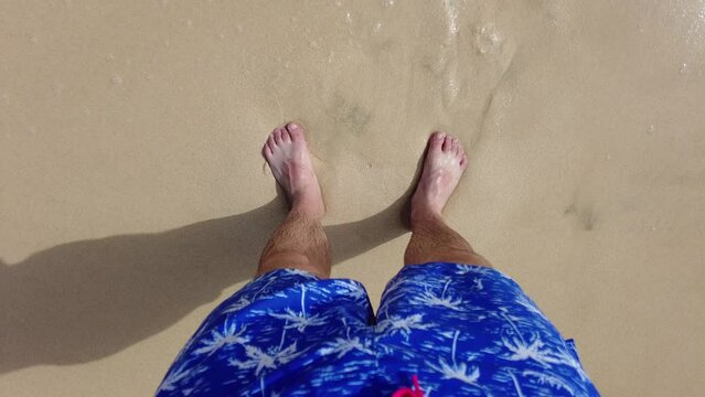 Top view of the legs of an adult man in blue beach shorts on a sandy shore with waves coming