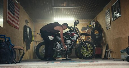 A male mechanic with a short haircut and a gray T-shirt puts his motorcycle on a jack in his garage workshop against of the US flag. Start of the working day and start of motorcycle maintenance in the