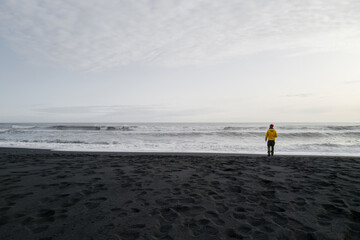 Obraz premium Man in a yellow jacket standing on the black sand beach overlooking the horizon