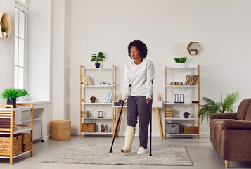 Young ethnic woman walking with crutches at home. African American girl with a broken leg walking...
