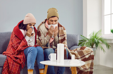 Warmly dressed man and woman having seasonal flu. Sick couple wrapped in blanket sitting on sofa in...