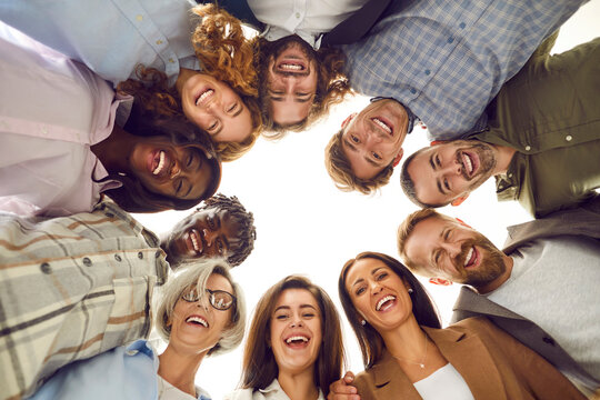 Happy diverse team having fun together. Low angle group portrait of cheerful joyful young and senior Caucasian and African American business people friends huddling, looking down at camera and smiling