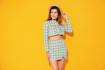 Young beautiful smiling female in trendy summer green costume clothes. Carefree woman posing near yellow wall in studio. Positive model having fun indoors. Cheerful and happy. Jacket and skirt, hat