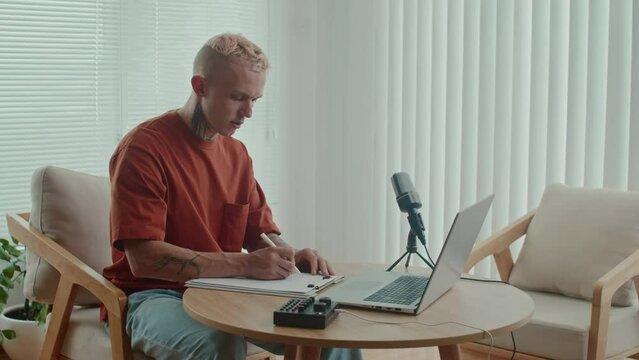 Medium long shot of concentrated male blogger working on script of his podcast while sitting in front of laptop and microphone