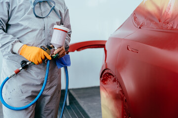 Unrecognizable automobile repairman worker painting a red car in a paint chamber during repair work.