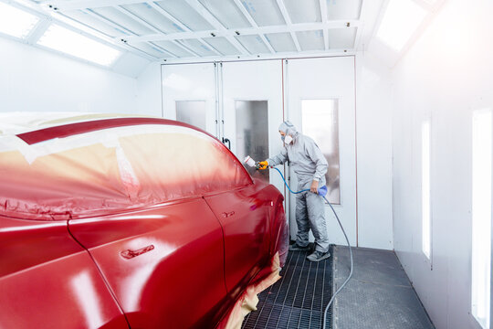 Wide angle of automobile repairman painter in protective workwear and respirator painting car body in paint chamber.
