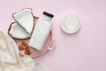 Pink ceramic plate with handle, a bottle of nut milk, walnuts, fresh coconut, milk glass and cloth towel on pastel background. Mockup with health and beauty theme