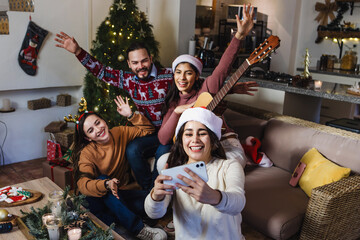 portrait of Hispanic friends taking a photo selfie with mobile phone sitting near Christmas tree at...