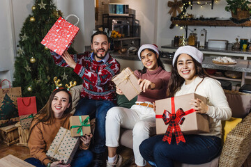 portrait of Hispanic friends holding gifts and sitting near Christmas tree at home in Mexico. Holidays and celebration concept in Latin America