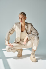 glamorous fashion model in beige suit and laced boots sitting on haunches in sunshine on grey
