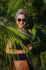 A girl in a colored turban, sunglasses with red lips on a background of greenery and palm leaves