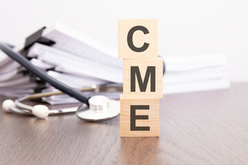text CME is written on wooden cube near a stethoscope on a dark background.