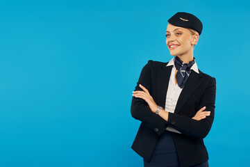 Obraz na płótnie Canvas joyful air hostess in corporate uniform posing with folded arms on blue backdrop, airline industry
