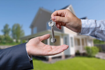 A hand giving a key to another hand. House in the background.