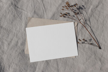 Winter minimal stationery. Blank Christmas greeting card, invitation mockup with dry flower and craft paper envelope in sunlight. Soft shadows. Linen tablecloth textured background. Flatlay, top view.