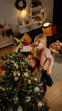 Mother lifting her little daughter so she can put a star on top of Christmas tree