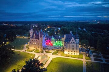 Moszna Castle from above at night: a fairytale like castle with many towers, illuminated by colorful spotlight, Polish Hogwarts. Poland.