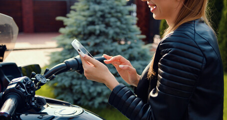 Fototapeta na wymiar Beautiful smiling trendy 20-aged brunette with high ponytail and black leather clothes browsing interesting apps on her mobile, posing on modern motorcycle.