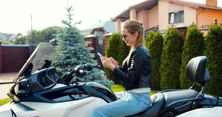 Smiling modern 20-aged woman biker with high ponytail and dressed in black leather clothes relaxing on luxurious white motorcycle on mototrack and writing sms on mobile phone.