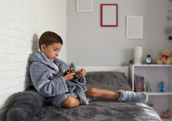 Cute 7 year old boy in gray robe and slippers sits on the bed.Boy plays with lego pieces. - obrazy, fototapety, plakaty