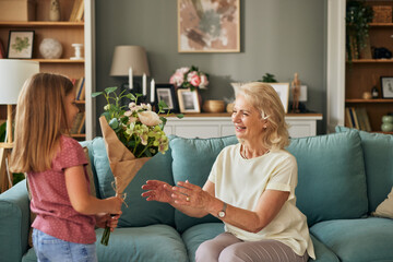 A young girl presents a bouquet of fresh flowers to her delighted grandmother in a cozy living room setting