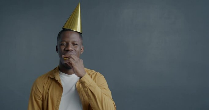 Slow motion portrait of joyful African American man wearing party hat blowing horn smiling looking at camera on gray background. Celebration and holiday concept.