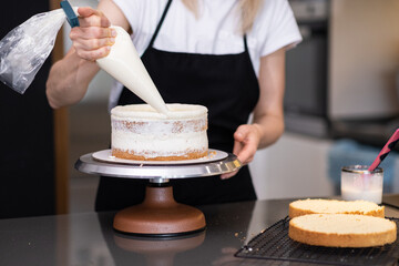 Skilled confectioner smearing white cream around sugar free cake layers with pastry cone standing at table woman in black apron making delicious confectionery for customer - obrazy, fototapety, plakaty
