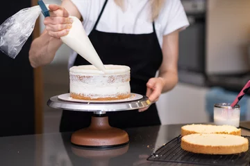 Tuinposter Skilled confectioner smearing white cream around sugar free cake layers with pastry cone standing at table woman in black apron making delicious confectionery for customer © olga_demina