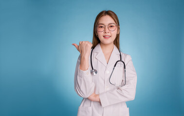 Young asian doctor wear medical uniform lab coat and stethoscope on blue background. Smiling female doctor pointing thumb to empty copy space for healthcare product or show banner rules safety.