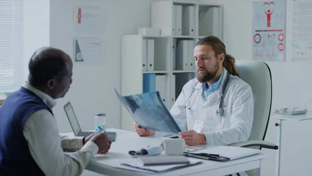 Medium shot of male Caucasian physician listening to African American patient and analyzing X-ray image during appointment in hospital